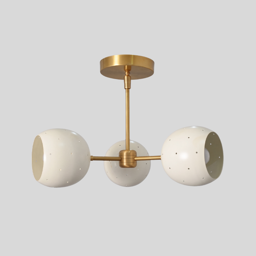 Triple white orb ceiling light featuring three perforated globe shades on a raw brass semi-flush mount, with a mid-century modern aesthetic and ambient pinhole lighting details.